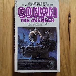 May include: A paperback book titled "Conan the Avenger" with a purple and white cover. The cover art depicts a muscular warrior in a battle scene with a dark background. The book is by Robert E. Howard, Bjorn Nyberg, and L. Sprague de Camp.
