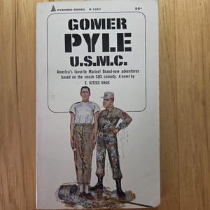 May include: A vintage paperback book titled "Gomer Pyle U.S.M.C." with a cover illustration of two men, one in uniform. The cover text reads "America's favourite Marine! Brand-new adventures based on the smash CBS comedy. A novel by E. KITZES KNOX."