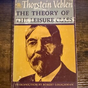 Puede incluir: Un libro de bolsillo titulado The Theory of the Leisure Class de Thorstein Veblen con una introducción de Robert Lekachman. La portada presenta el retrato de un hombre con barba sobre un fondo amarillo, con bordes y texto en color morado.