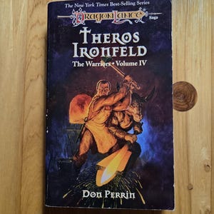 May include: A paperback book titled "Theros Ironfeld" from the Dragonlance Saga series. The cover features a warrior figure striking a horned creature with a hammer. The author's name, Don Perrin, is at the bottom.