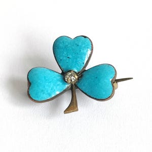 May include: A vintage shamrock brooch with three turquoise enamel leaves and a central clear rhinestone. The brooch has a gold-tone metal frame and pin, and is set against a white background.