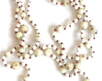Puede incluir: Un collar largo que presenta un patrón repetitivo de pequeñas cuentas blancas lisas y cuentas más grandes, redondas y de color blanco roto con un diseño tallado de estrella en color marrón oscuro. Las cuentas están ensartadas en un cordón fino rojizo.