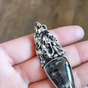 Puede incluir: Un colgante de plata con una secci&oacute;n superior de metal texturizado y org&aacute;nico con un aspecto irregular y fundido. La parte inferior contiene una piedra triangular transl&uacute;cida con inclusiones oscuras y una fina banda amarilla.