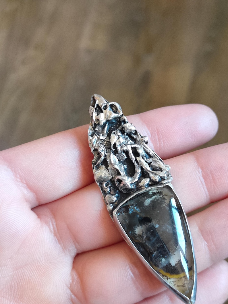 Puede incluir: Un colgante de plata con una secci&oacute;n superior de metal texturizado y org&aacute;nico con un aspecto irregular y fundido. La parte inferior contiene una piedra triangular transl&uacute;cida con inclusiones oscuras y una fina banda amarilla.