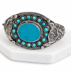 May include: A silver cuff bracelet featuring a large central oval turquoise stone surrounded by a ring of fourteen smaller round turquoise cabochons. The metal surface displays intricate embossed floral patterns and twisted rope detailing.