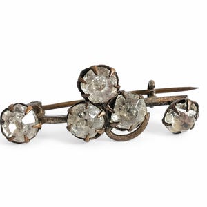 May include: A brooch featuring four faceted, clear, round stones set in a dark, oxidized metal frame. The stones are arranged in a cluster with a pin mechanism visible on the back. The metal has a textured, aged appearance with prong settings.