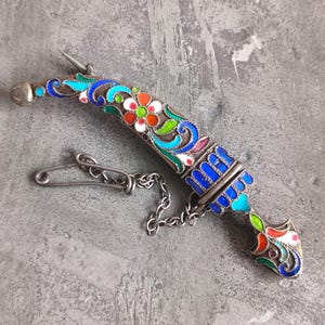 Könnte beinhalten: Ein dekorativer Dolch mit silberner Klinge und Griff. Der Griff ist mit bunten Emaille-Blumendesigns in Rot, Blau, Grün und Weiß verziert. Eine Silberkette und eine Sicherheitsnadel sind am Griff befestigt.
