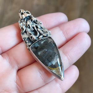 Puede incluir: Un colgante de plata con una piedra preciosa triangular de color gris oscuro engastada en un marco de plata texturizado y detallado.