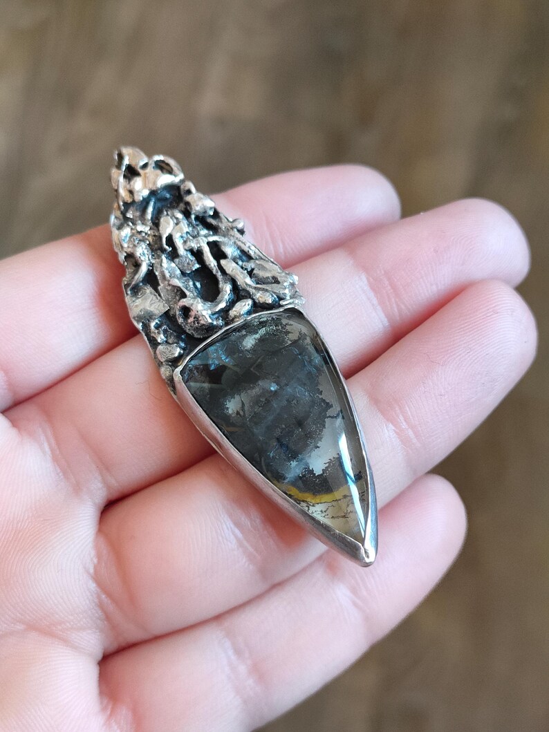 Puede incluir: Un colgante de plata con una piedra preciosa triangular de color gris oscuro engastada en un marco de plata texturizado y detallado.