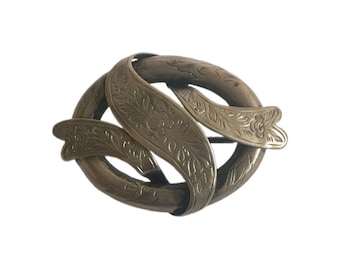 May include: An oval-shaped brooch crafted from a metallic material with a muted, aged finish. The design features an intertwined, ribbon-like structure decorated with delicate, engraved floral and leaf patterns across its entire surface.