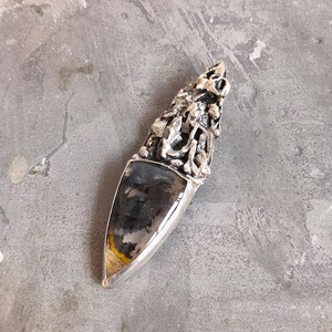 Puede incluir: Un colgante de plata con una piedra negra y dorada engastada en una forma puntiaguda. La plata est&aacute; dise&ntilde;ada de forma intrincada con un patr&oacute;n texturizado y org&aacute;nico.