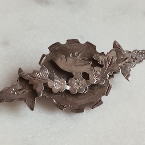Pode incluir: Broche prateado com um pássaro pousado em um design floral e de folhas. O broche tem um centro circular com uma borda dentada, e o pássaro é cercado por flores e folhas delicadas.