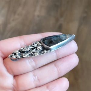 Puede incluir: Un colgante de plata con un dise&ntilde;o texturizado org&aacute;nico y una piedra preciosa gris oscuro engastada en el centro. El colgante tiene un bucle para sujetar una cadena.