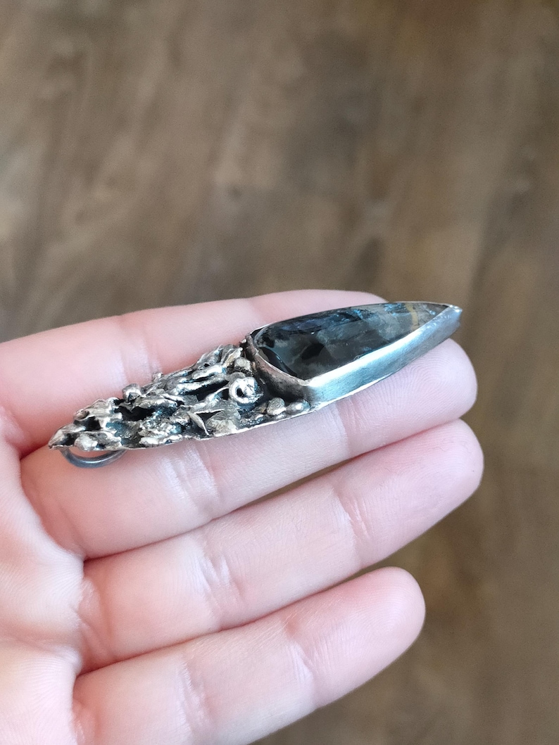Puede incluir: Un colgante de plata con un dise&ntilde;o texturizado org&aacute;nico y una piedra preciosa gris oscuro engastada en el centro. El colgante tiene un bucle para sujetar una cadena.