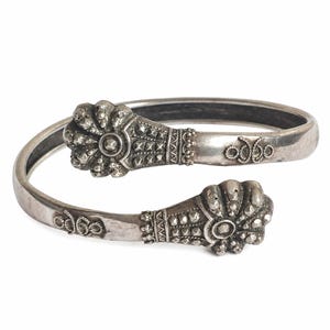 Pode incluir: Uma pulseira de prata tipo bypass com dois terminais ornamentados em forma de leque, decorados com pequenas pedras facetadas. A banda apresenta um trabalho de metal intrincado, incluindo tachas piramidais texturizadas e detalhes em corda.