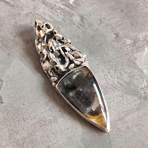 May include: A silver pendant featuring a dark grey and yellow agate stone. The silver is intricately designed with a textured, organic pattern.