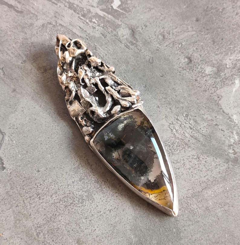 May include: A silver pendant featuring a dark grey and yellow agate stone. The silver is intricately designed with a textured, organic pattern.