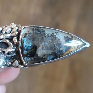 Puede incluir: Un anillo de plata con una piedra triangular transl&uacute;cida que presenta inclusiones oscuras con forma de &aacute;rbol dendr&iacute;tico. El engaste de metal tiene una superficie texturizada, org&aacute;nica e irregular que rodea la base de la piedra.