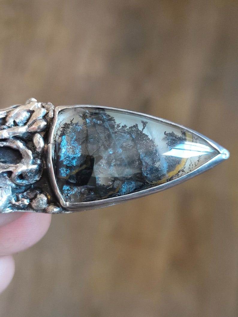 Puede incluir: Un anillo de plata con una piedra triangular transl&uacute;cida que presenta inclusiones oscuras con forma de &aacute;rbol dendr&iacute;tico. El engaste de metal tiene una superficie texturizada, org&aacute;nica e irregular que rodea la base de la piedra.