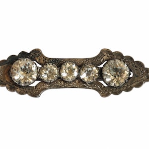 Pode incluir: Um broche de metal prateado de formato alongado com uma borda festonada texturizada e uma fileira de cinco pedras redondas transparentes facetadas. As duas pedras externas são maiores que as três pedras centrais menores.