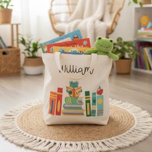 May include: A cream-colored canvas tote bag featuring the name William in black script. The bag displays a colorful graphic of a green frog reading a book atop a stack of books, with additional books and a red apple. A plush green frog sits on the rim.