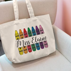 May include: White canvas tote bag with a rainbow of crayons and the text "Miss Elaine" printed on the front.