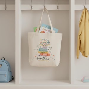 May include: A cream-colored tote bag with a colorful illustration of stacked books, a rainbow, and clouds. The name "Jonathan" is written in cursive below the illustration. The bag is filled with books and hangs from a metal hook.