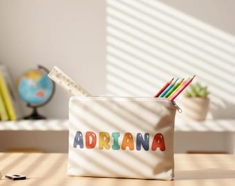 May include: A beige canvas pencil case with the name "ADRIANA" in colorful bubble letters. The case contains a ruler and several colored pencils. The pencil case is sitting on a wooden surface.