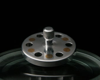 May include: A silver spinning top with a textured top and gold accents. The top is spinning on a clear glass surface.