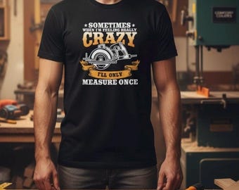 Op de afbeelding: Zwart t-shirt met de tekst "Sometimes When I'm Feeling Really Crazy I'll Only Measure Once" en een afbeelding van een cirkelzaag. De persoon draagt het shirt in een werkplaats.