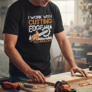 May include: Black t-shirt featuring the text "I WORK WITH CUTTING EDGE TECHNOLOGY" in orange and white, with a chainsaw graphic. The image shows the shirt being worn in a workshop environment.