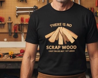 Op de afbeelding: Zwart t-shirt met de tekst "THERE IS NO SCRAP WOOD (JUST PIECES NOT YET USED)" en een afbeelding van houten planken. Het t-shirt wordt gedragen door een persoon in een werkplaats.