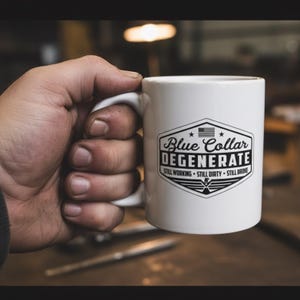 Könnte beinhalten: Weiße Keramik-Tasse mit dem Text "Blue Collar Degenerate" in einem schwarz-weißen Design. Die Tasse trägt auch den Text "Still Working, Still Dirty, Still Broke". Die Tasse wird von einer Hand gehalten.