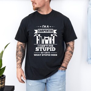 May include: Black t-shirt with a white graphic that reads "I'M A CARPENTER, I CAN'T FIX STUPID, BUT I CAN FIX WHAT STUPID DOES." The design includes hammer and saw illustrations.