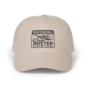 May include: A beige baseball cap with a curved brim. The front features a black and white patch that reads "PROFESSIONAL Crop DUSTER SILENT BUT DEADLY" with an illustration of a biplane. The cap is made of a soft fabric.