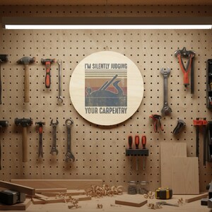 May include: A round wooden sign with the text "I'M SILENTLY JUDGING YOUR CARPENTRY" and a graphic of a hand plane. The sign is hanging on a pegboard wall with various carpentry tools, including hammers, wrenches, and pliers.