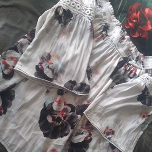 May include: White off-the-shoulder blouse with a floral print featuring black, pink, and blue flowers. The blouse has a lace trim around the neckline and sleeves.