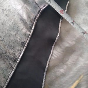 May include: Close-up of a silver fabric with a black fabric seam. A measuring tape is visible with the numbers 30, 31, 32, 33, 34, and 35 inches.