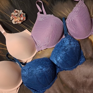 May include: A collection of four bras featuring two smooth, solid light peach bras and two textured, lace-covered bras in lavender and deep blue. Each bra includes adjustable shoulder straps and molded cups, showcasing a variety of fabric finishes.