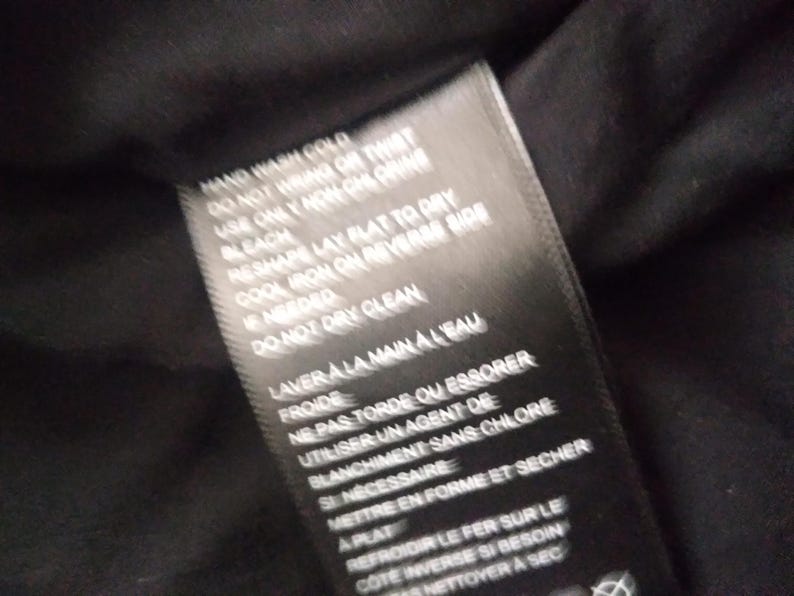 May include: Black care label with white text detailing washing instructions. Hand wash cold, do not wring or twist, use only non-chlorine bleach, reshape lay flat to dry, cool iron on reverse side if needed, do not dry clean, laver a la main a l'eau froide, ne pas tordre ou essorer, utiliser un agent de blanchiment sans chlore si necessaire, mettre en forme et secher a plat, refroidir le fer sur le cote inverse si besoin, nettoyer a sec.