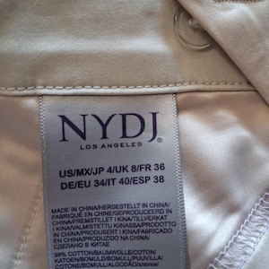 May include: Close-up of a light beige garment label from NYDJ Los Angeles. The label displays size information and fabric composition: 98% cotton and 2% elastane. The garment is made in China.