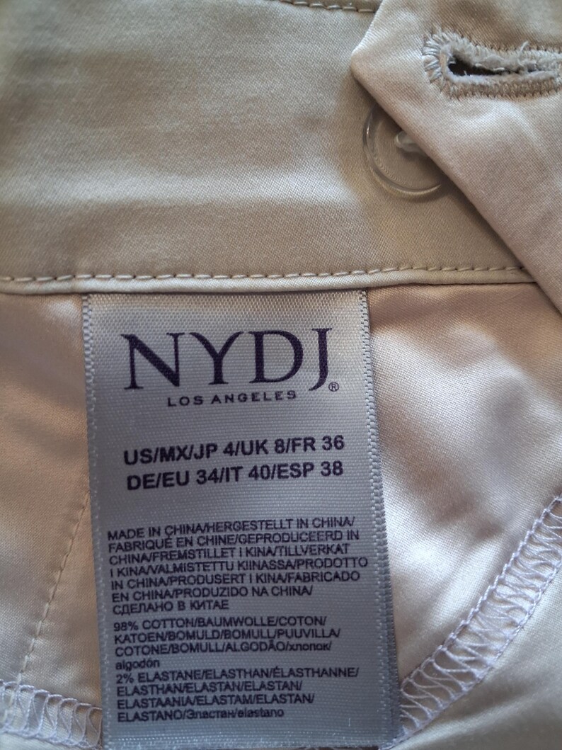 May include: Close-up of a light beige garment label from NYDJ Los Angeles. The label displays size information and fabric composition: 98% cotton and 2% elastane. The garment is made in China.