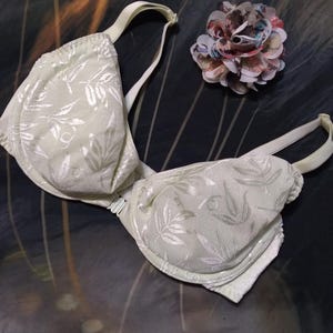 May include: A light green bra with a leaf pattern and white straps. A decorative flower made of colorful fabric is next to the bra. The bra has a classic design.