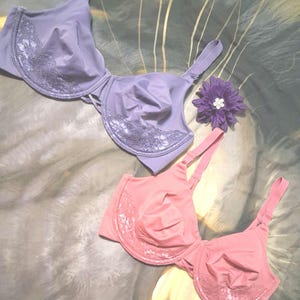 May include: A lavender underwire bra and a dusty rose underwire bra, both featuring lace detailing on the lower cups and adjustable shoulder straps, accompanied by a single purple fabric flower with a small white center embellishment.