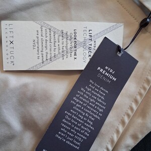 May include: Close-up of two tags from NYDJ Premium Denim. One tag reads "LIFT TUCK TECHNOLOGY" and the other says "NYDJ PREMIUM DENIM". The tags are black and white with text describing the denim's features and technology.