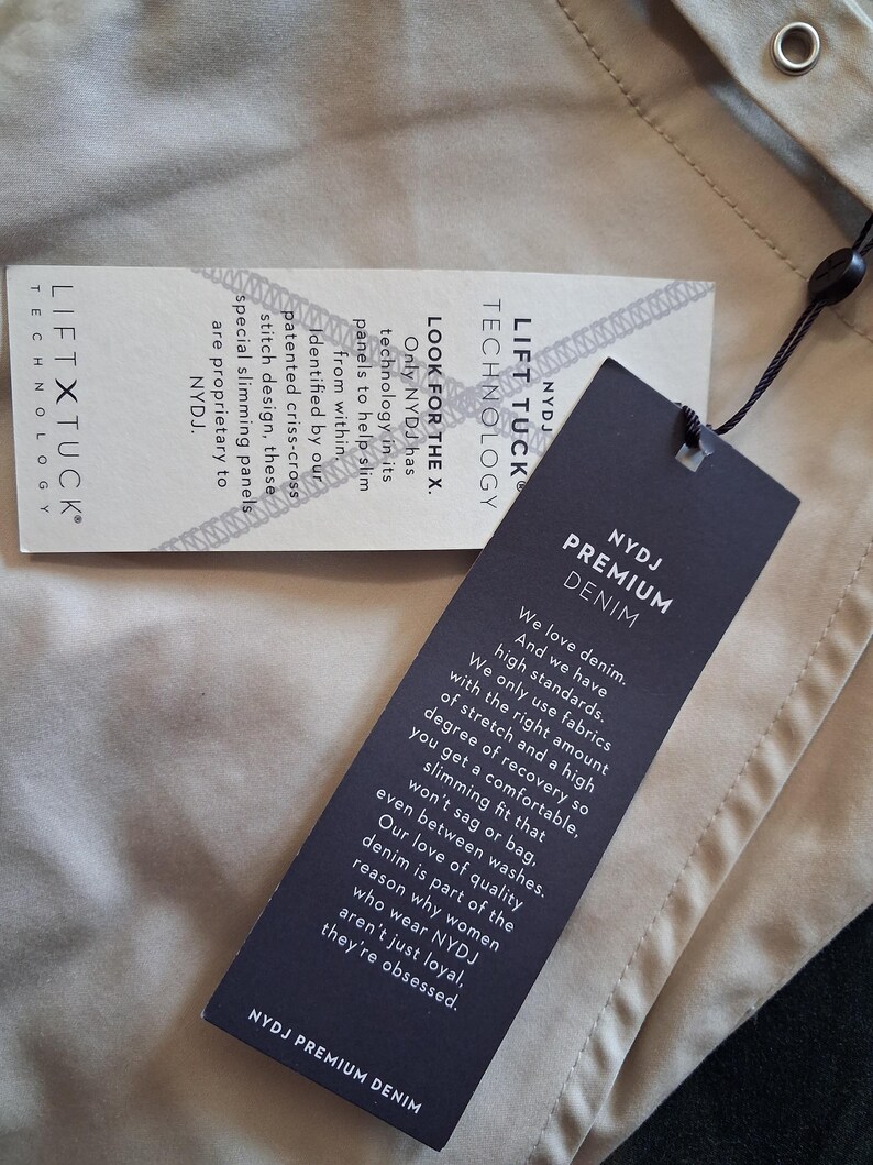 May include: Close-up of two tags from NYDJ Premium Denim. One tag reads "LIFT TUCK TECHNOLOGY" and the other says "NYDJ PREMIUM DENIM". The tags are black and white with text describing the denim's features and technology.