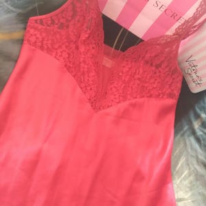 May include: Coral-colored satin slip with lace detailing on the bodice and straps. The lingerie features a V-neck design and delicate lace trim. A pink and white striped shopping bag is in the background.
