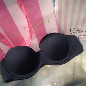May include: A strapless, navy blue bra is displayed in front of two shopping bags. The bags are pink and white striped, with the words "Victoria's Secret" printed on them in gold and black.