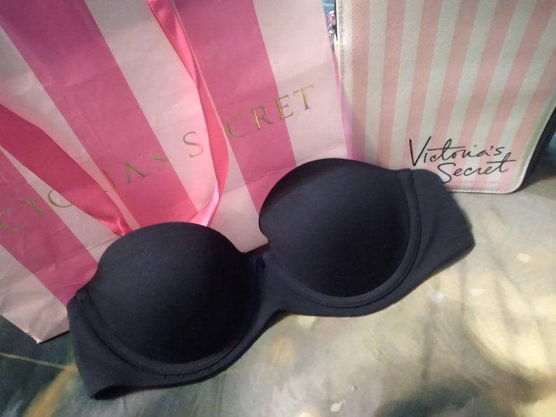 May include: A strapless, navy blue bra is displayed in front of two shopping bags. The bags are pink and white striped, with the words "Victoria's Secret" printed on them in gold and black.