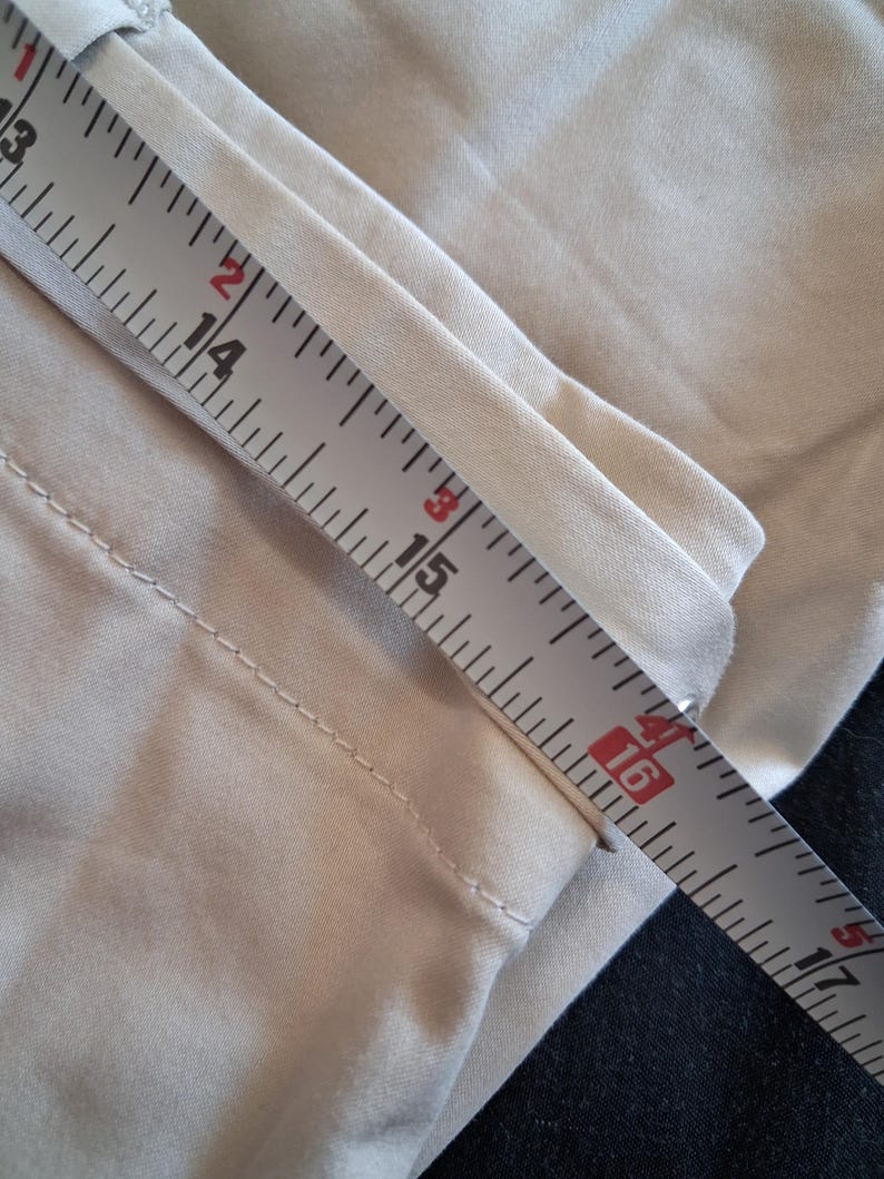 May include: Close-up of tan fabric being measured with a ruler. The ruler shows measurements from 13 to 17 inches. The fabric appears to be part of a garment, possibly pants or a skirt. The fabric is a solid, neutral color.